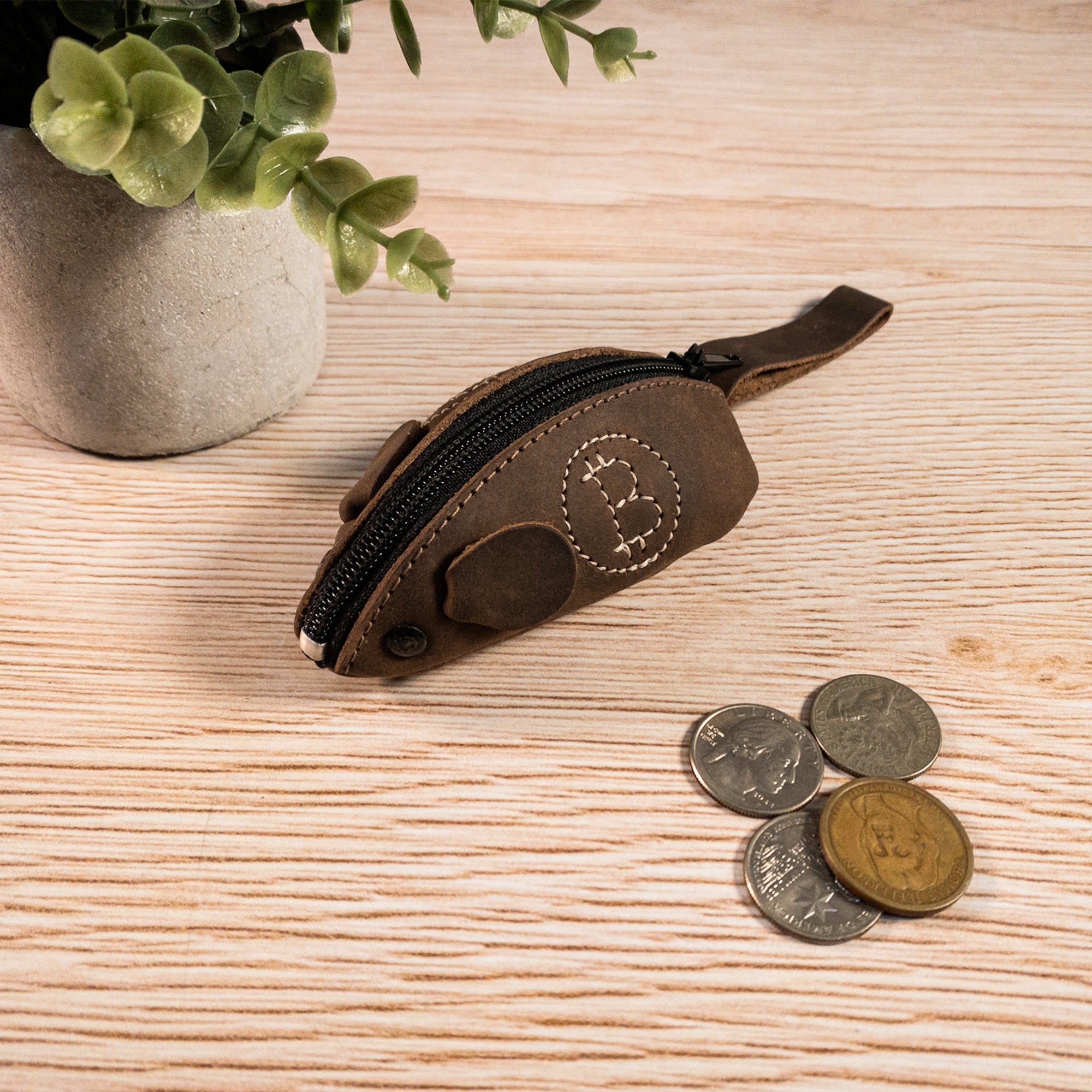 The Mouse Coin Pouch by Hide & Drink displayed on a wooden surface, next to various coins, illustrates its charming design. Made of full grain leather, this practical mouse-shaped change pouch serves as an ideal pocket case for coins, blending style with functionality. Handcrafted in La Antigua Guatemala and Oaxaca, Mexico, it reflects the beauty of handmade artisan products.