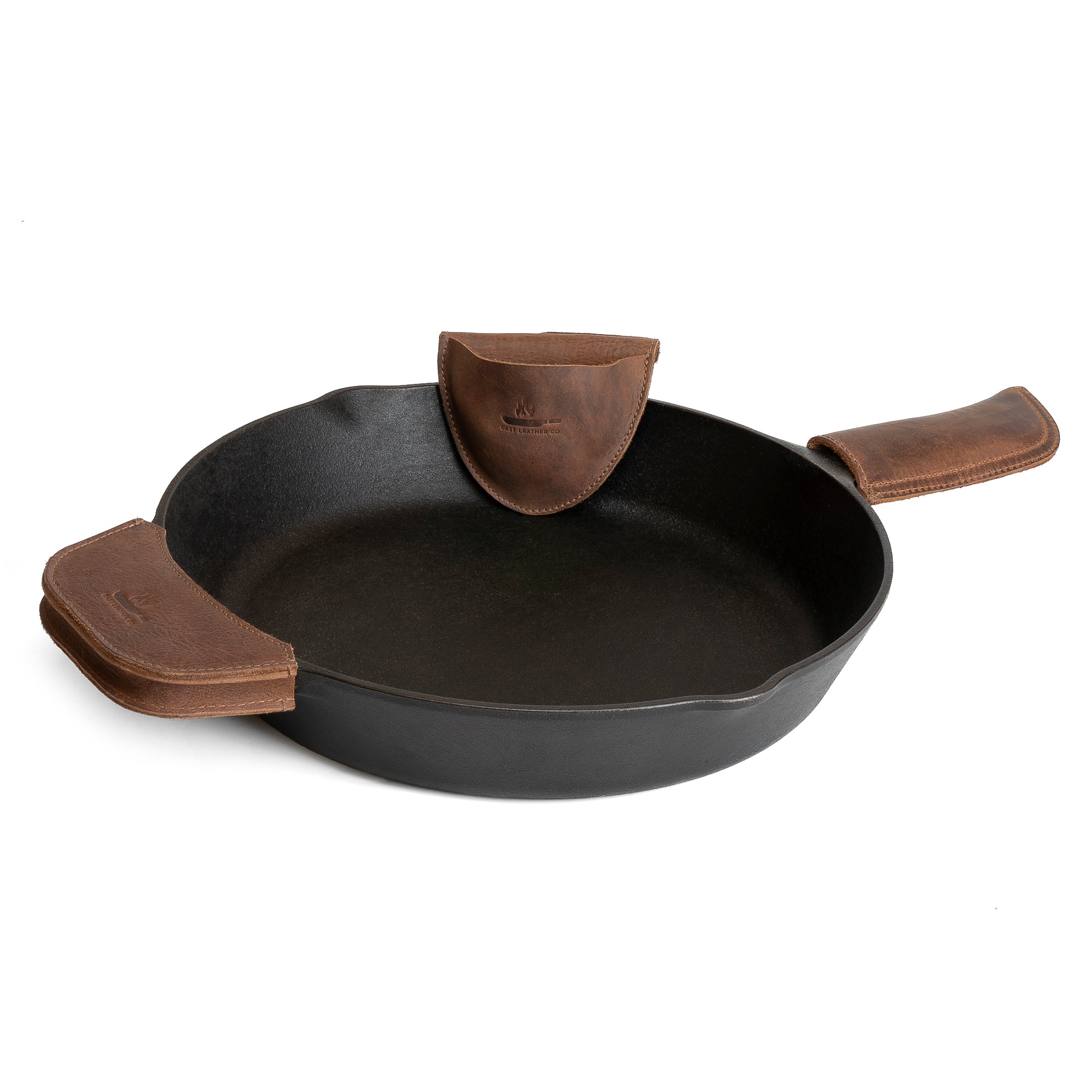 Showcasing the Hot Handle Holders Set of 3 by Cast Leather Co., this image features a cast iron pan equipped with rustic grip holders made from full grain leather. These stylish and functional additions serve as a panhandle, side kick, and grip mitt, providing an assist grip for cast iron skillets and pans. The handmade leather pieces are crafted through small batch production, ensuring quality and tradition, all made in La Antigua Guatemala & Oaxaca Mexico.
