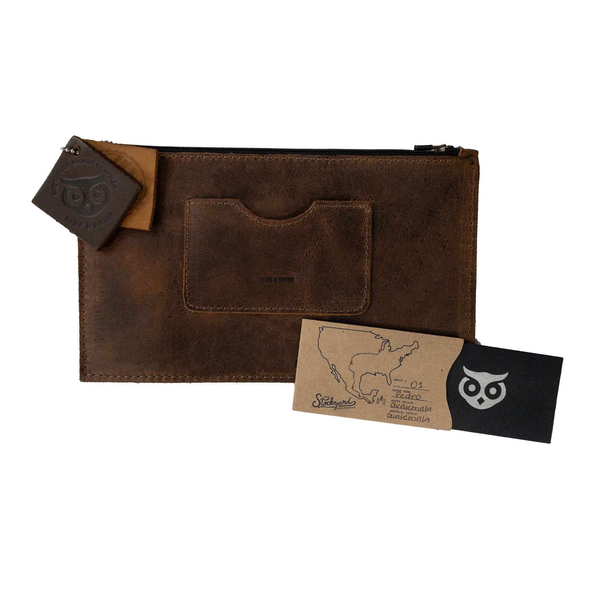 A brown rectangular zippered handbag by Hide & Drink alongside leather care tags, perfect for women with its card slot. This female purse, handmade from full grain leather, highlights artisan dedication with small batch production, showcasing craftsmanship that's fire branded and designed in La Antigua Guatemala & Oaxaca Mexico.