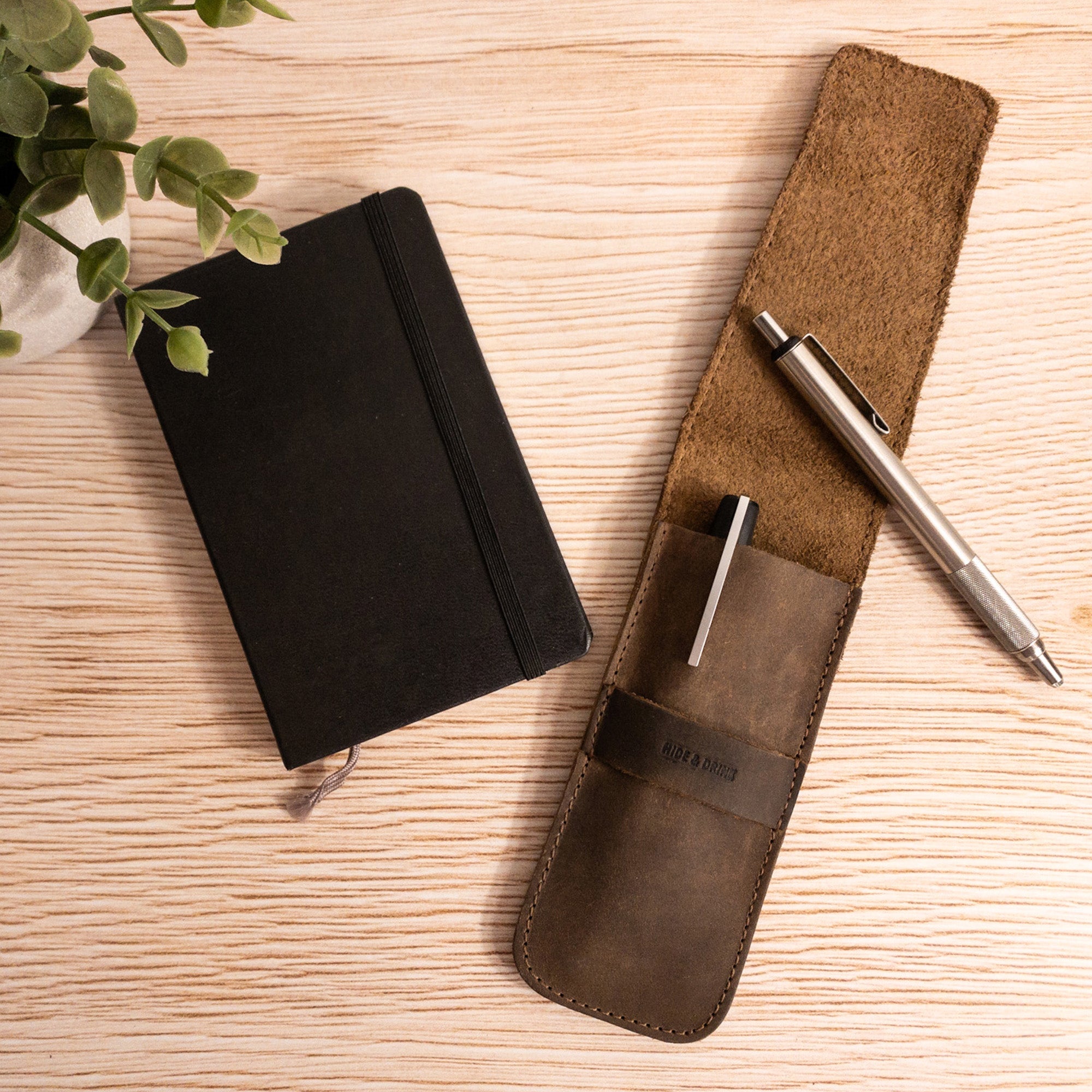 A styled image of the Hide & Drink Pen Case paired with a notebook and pen, presenting a functional office setup. This Pen Case Holder functions well as a Multi Pen Pouch to organize your essentials. Made from robust Full Grain Leather, this piece demonstrates StockyardX's commitment to small batch production. Hand cut and hand sewn craftsmanship shines through, featuring a fire branded logo - truly an artisan creation, handmade in La Antigua Guatemala & Oaxaca Mexico.