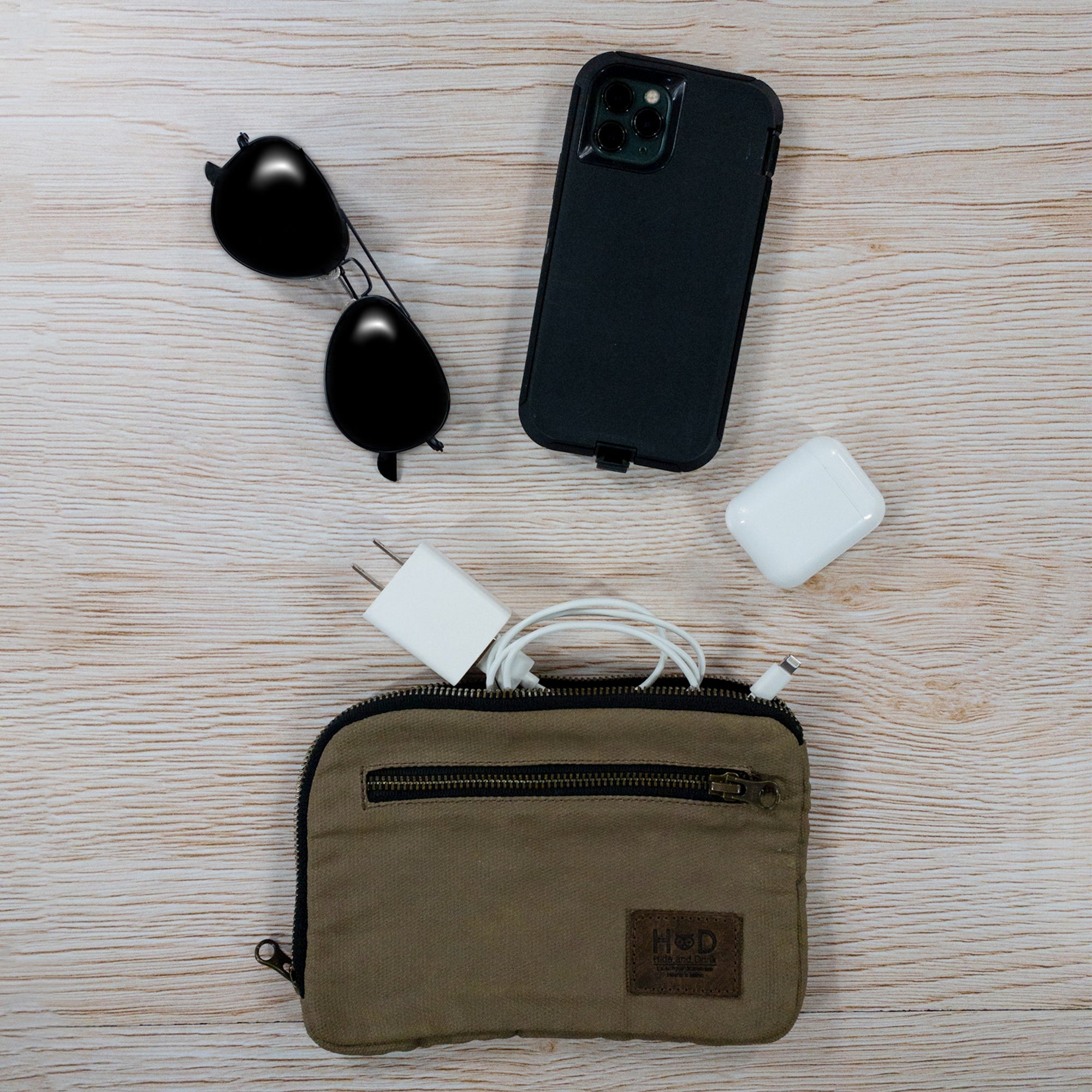 Hide & Drink's Zipper Gear Pouch alongside sunglasses and tech accessories, showcasing its utility as a travel bag crafted from water resistant waxed canvas.