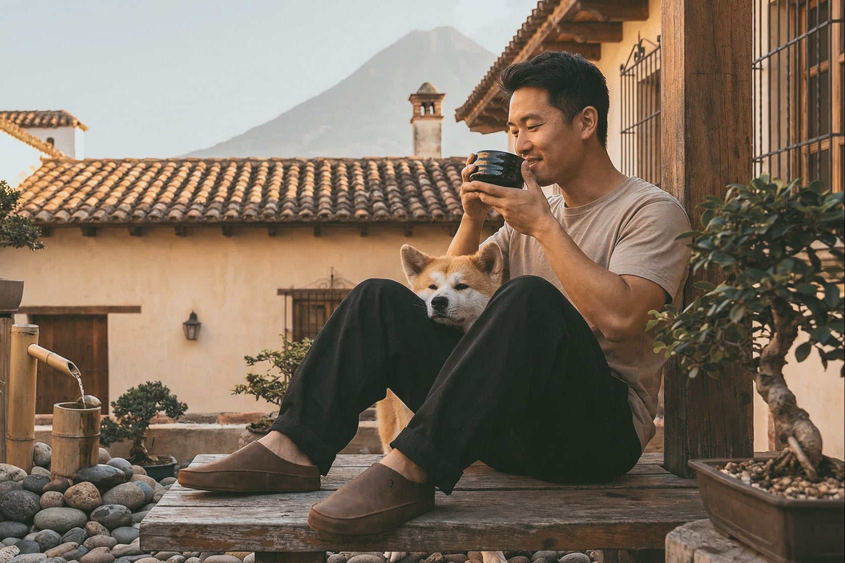 Man sitting on a wooden bench with a dog, holding a coffee cup, in a scenic outdoor setting.