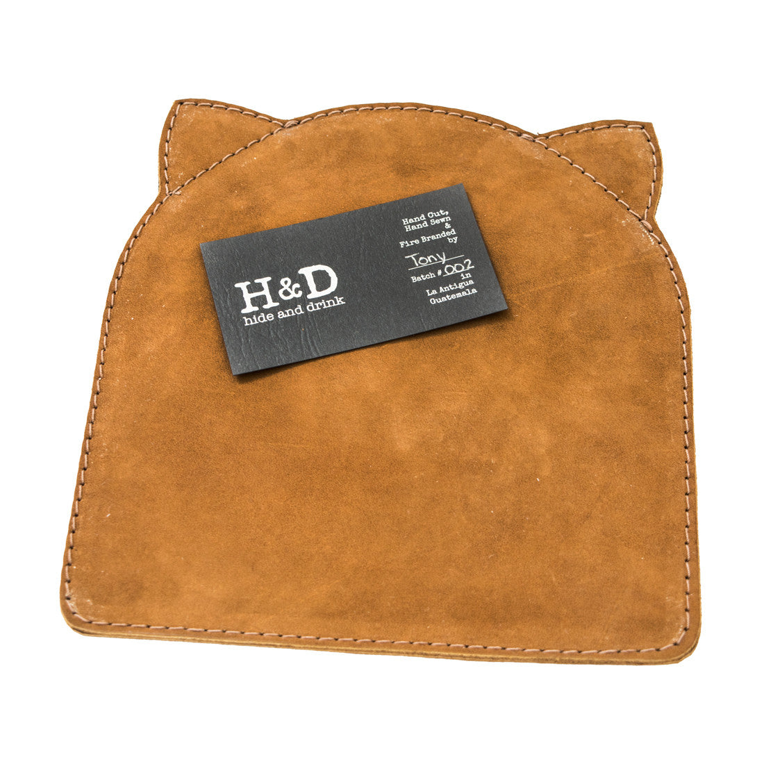 Soft Leather Cat Mouse Pad by Hide & Drink, displaying its unique cat ear shape and a business card for authenticity. Made from full grain leather, this artisan piece features small batch production, hand cut and hand sewn techniques, fire branded details, and is handmade in La Antigua Guatemala & Oaxaca Mexico.