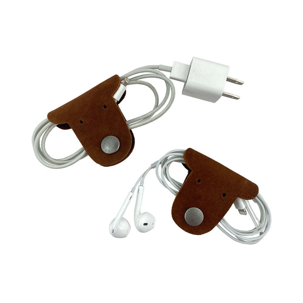 Two brown Dog Shaped Cord Keepers holding charging cables and earbuds, an adorable way to keep cords organized by Hide & Drink. Dog Shaped Cord Keeper (Cord Clam) 2-Pack, Full Grain Leather, StockyardX, Small Batch Production, Hand Cut, Hand Sewn, Fire Branded, Artisan, Handmade in La Antigua Guatemala & Oaxaca Mexico.