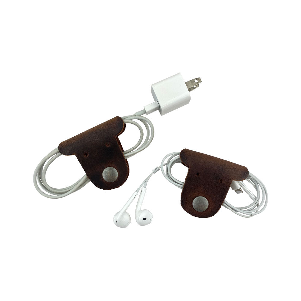 Two dark brown Dog Shaped Cord Keepers neatly organizing earbuds and charging cords, an adorable design by Hide & Drink for stylish storage. Dog Shaped Cord Keeper (Cord Clam) 2-Pack, Full Grain Leather, StockyardX, Small Batch Production, Hand Cut, Hand Sewn, Fire Branded, Artisan, Handmade in La Antigua Guatemala & Oaxaca Mexico.