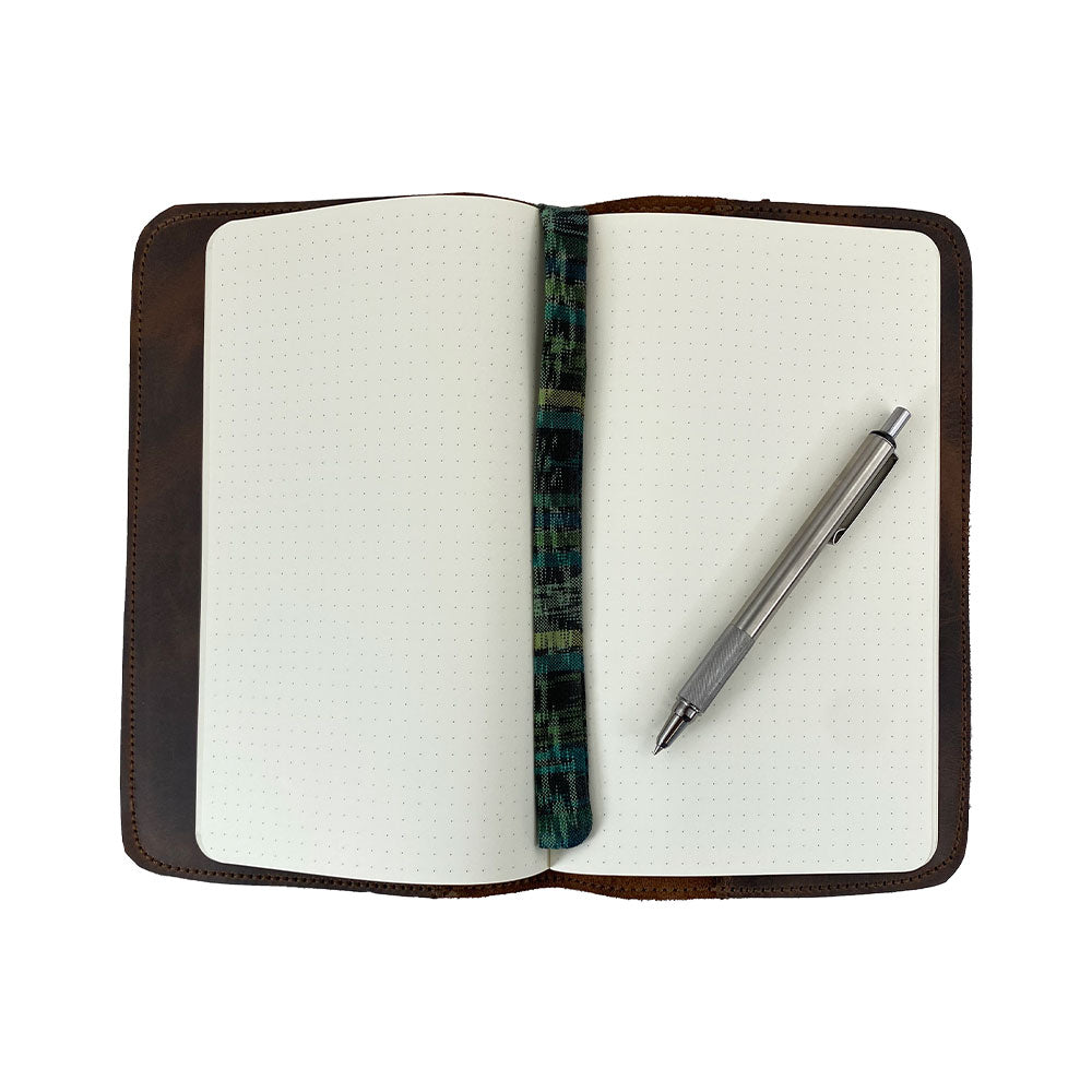 Opened Journal Cover for Moleskine Cahier L (5 x 8.25 in.) with a pen resting on blank pages by Hide & Drink. This refillable journal cover with a strap features rustic full grain leather, perfect for large size (5 x 8.25 in.) notebooks, showcasing the artisan craftsmanship.