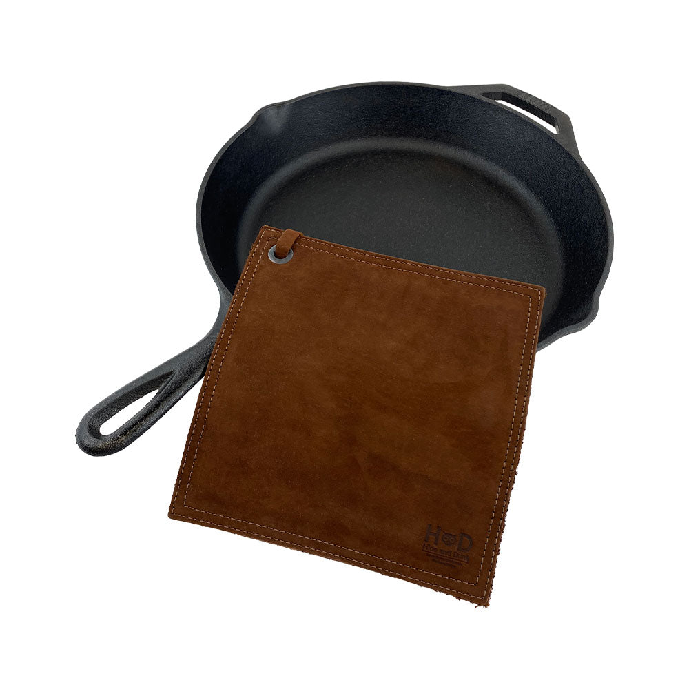Hide & Drink Pot Pad beside a cast iron pan, demonstrating its utility. This rustic hot pot pad is double-layered with full grain leather, making it an essential kitchen tool. Handmade in La Antigua Guatemala & Oaxaca Mexico.
