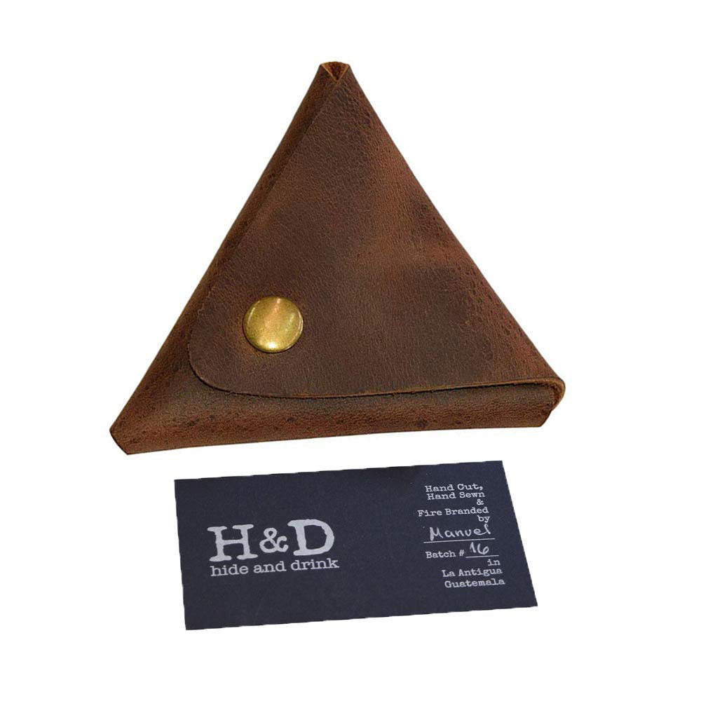 The Triangle Coin Pouch by Hide & Drink displayed next to its informational card, highlighting its handmade nature. This Triangle Coin Pouch is made from exquisite Full Grain Leather, suitable for holding cash and cords. Celebrate artisan craftsmanship and small batch production with this handmade item, which is fire branded and crafted in La Antigua Guatemala & Oaxaca Mexico.