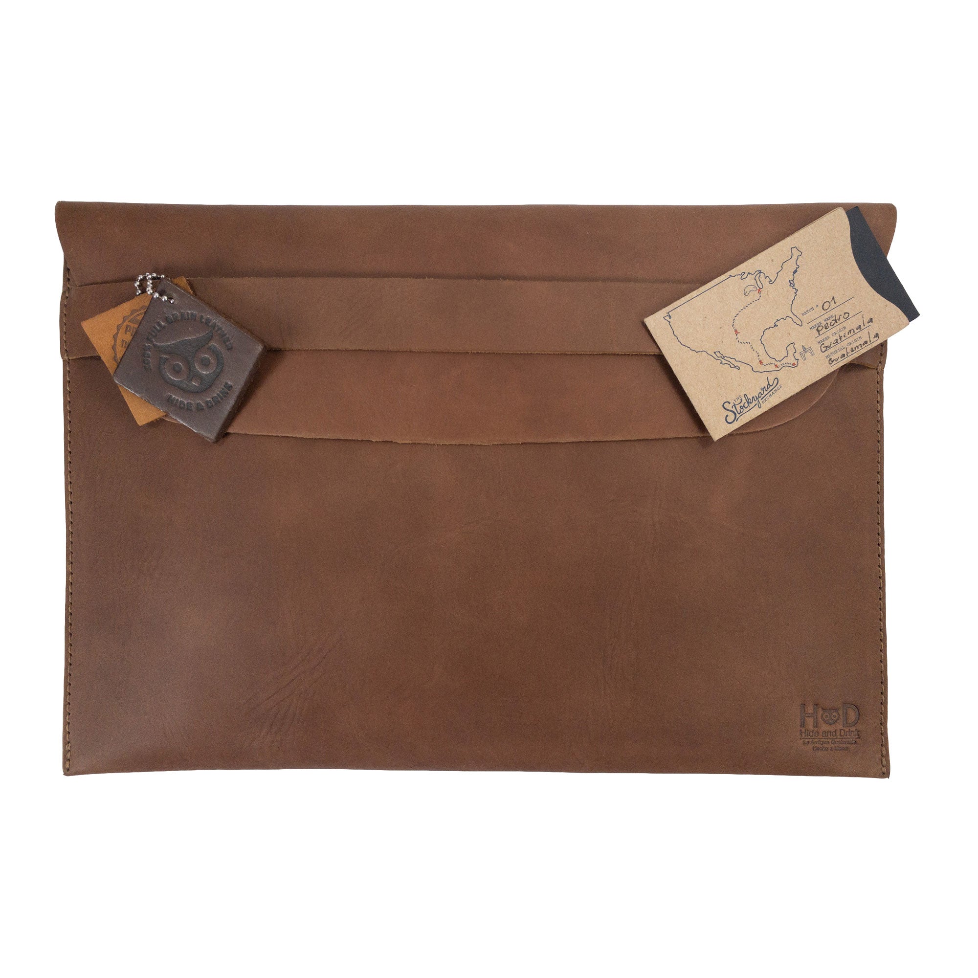 Front view of the brown Hide & Drink MacBook Slim Case with decorative tags, showcasing its rustic appeal. This case is made for MacBook Air/Pro 13 Inch with M1 Chip, emphasizing the artisan's work with hand cut and hand sewn designs.