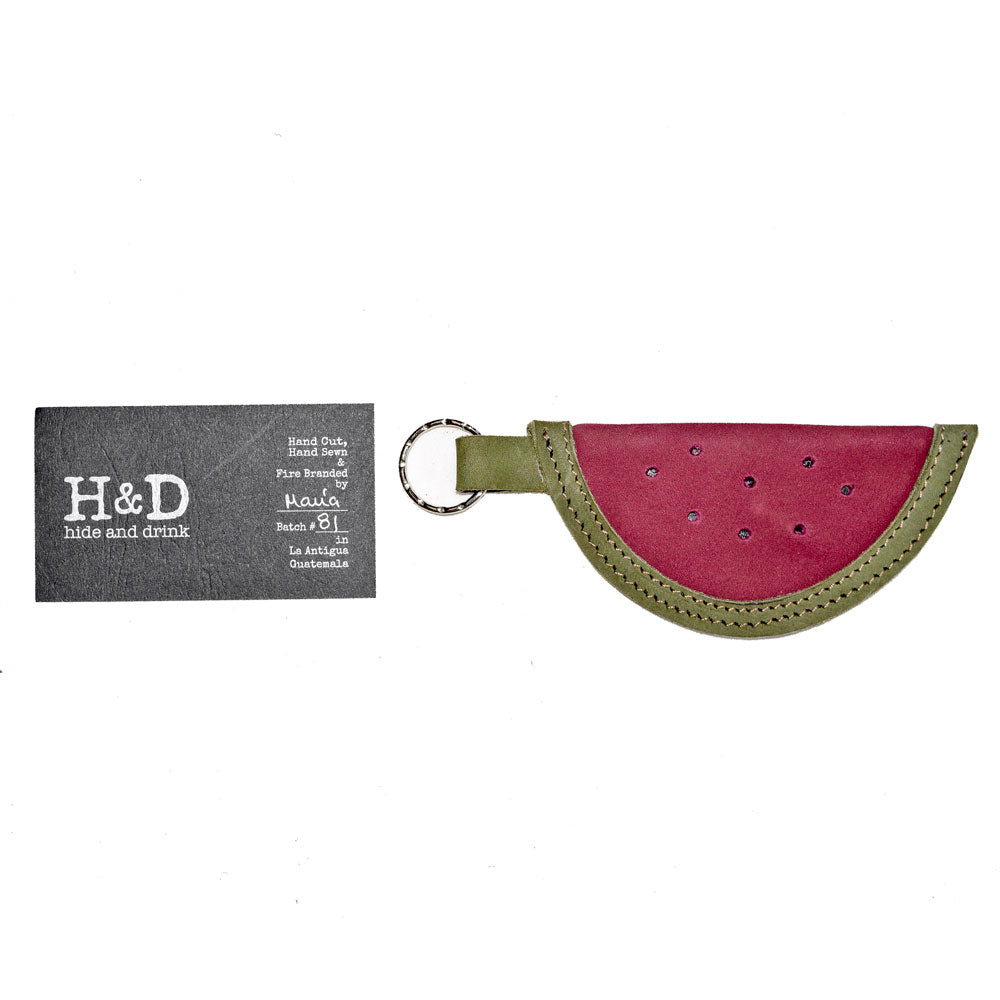 The Watermelon Keychain by Hide & Drink alongside its packaging, radiating charm and character. This delightful key holder is a product of small batch production techniques utilizing premium full grain leather, representing artisan craftsmanship. A beautiful blend of function and style, handmade in La Antigua Guatemala & Oaxaca, Mexico.