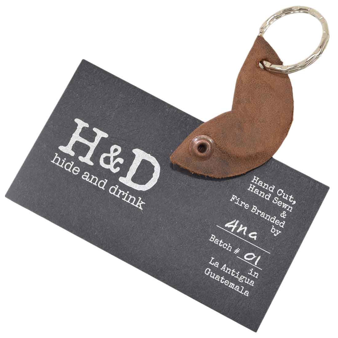 The Fidget Cashew Keychain by Hide & Drink is displayed alongside a business card, accentuating its stylish design and artisan quality. This key holder stands as a Good Luck Charm made from durable Full Grain Leather, demonstrating the small batch production technique that includes hand cut, hand sewn, and fire branded craftsmanship from La Antigua Guatemala and Oaxaca Mexico.