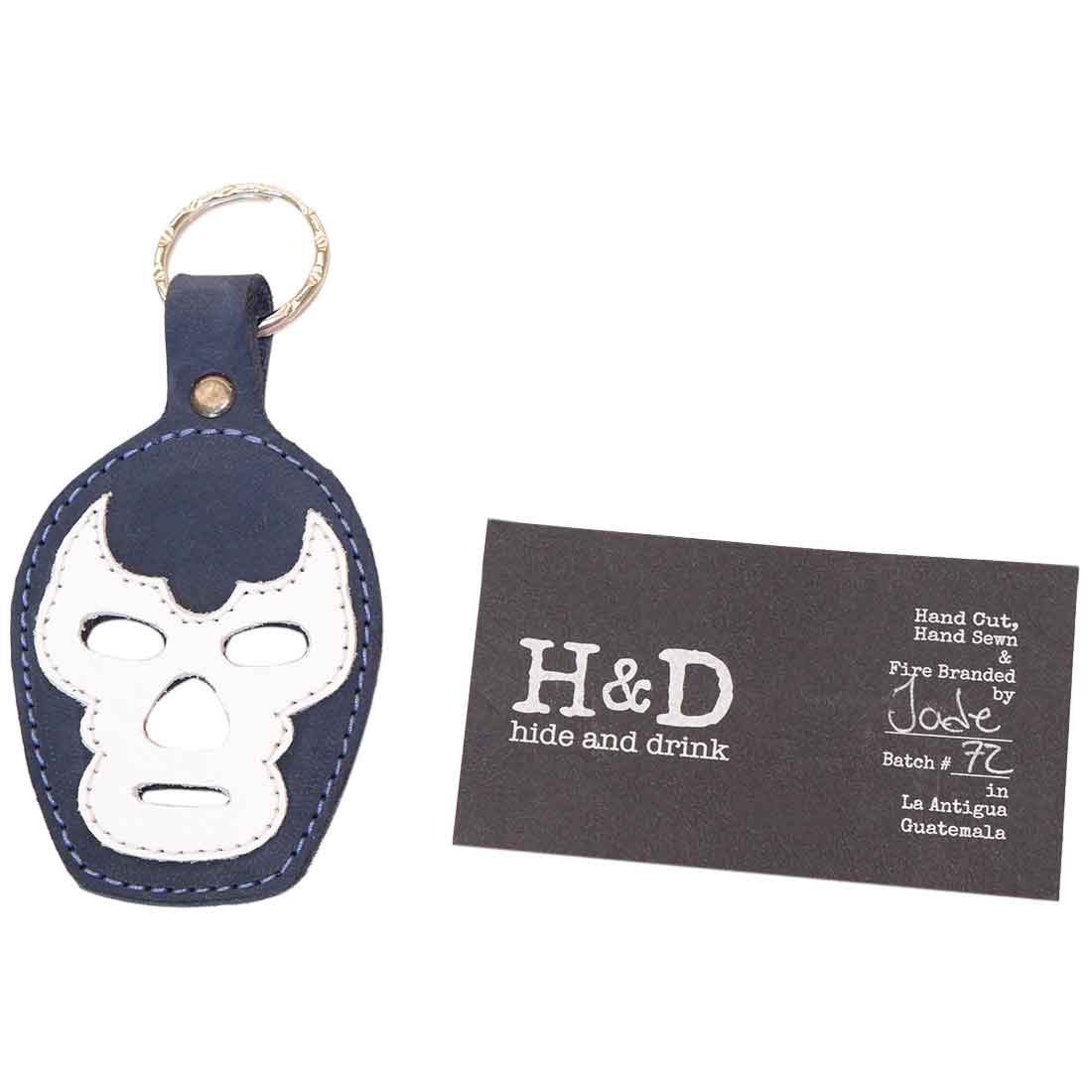 The Luchador Mask Leather Keychain with its brand card, highlighting the craftsmanship behind this unique accessory. Made by Hide & Drink, this keyring is a stylish key organizer crafted from full grain leather, showcasing artisan qualities of hand cut and hand sewn finishes, handmade in La Antigua Guatemala and Oaxaca Mexico.