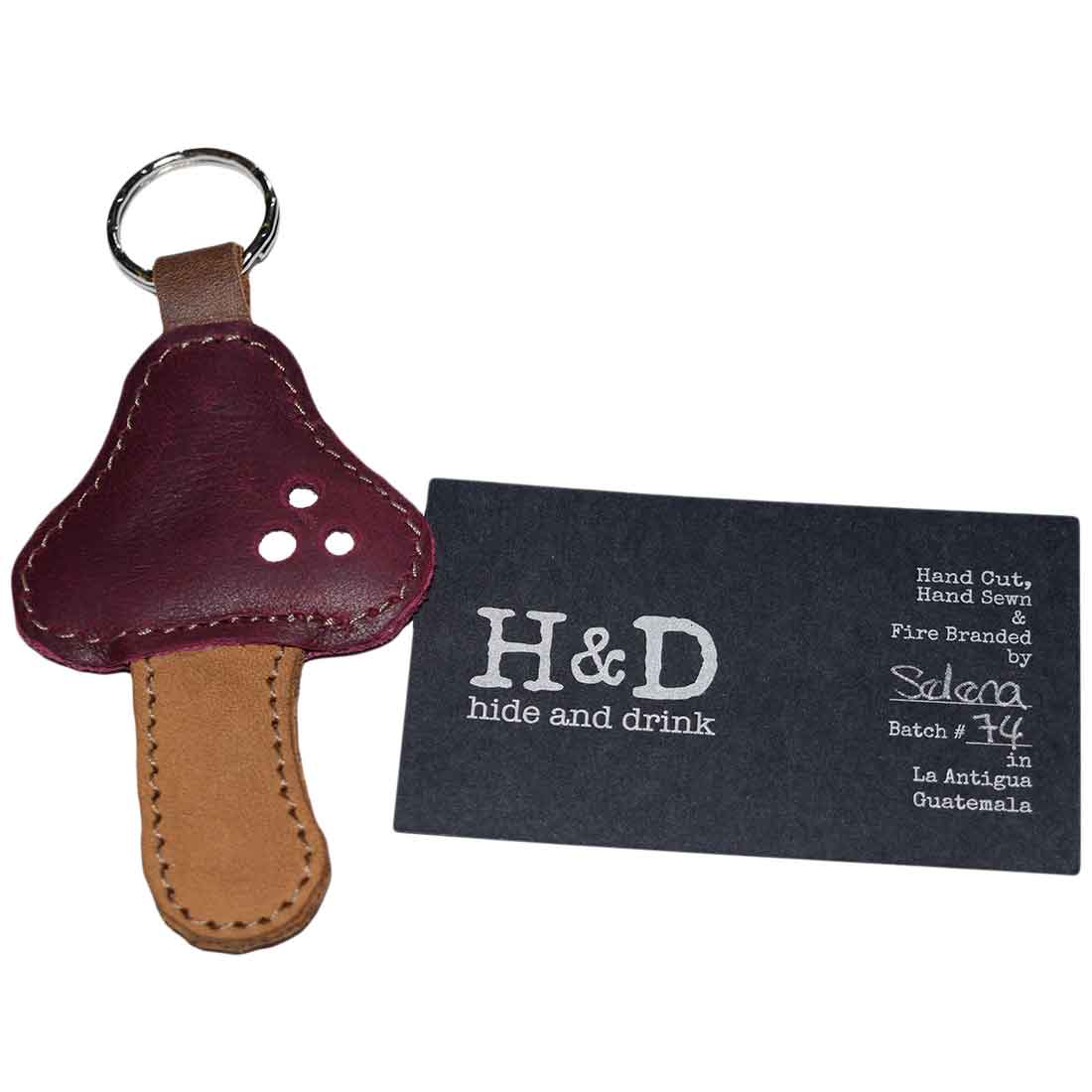 The Red Mushroom Keychain by Hide & Drink displayed alongside its branding card, emphasizing its quality and craftsmanship. Made of Full Grain Leather, this keychain is a standout accessory reflecting StockyardX's small batch production methods. Hand cut, hand sewn, and fire branded, it represents artisan work made in La Antigua Guatemala & Oaxaca Mexico.