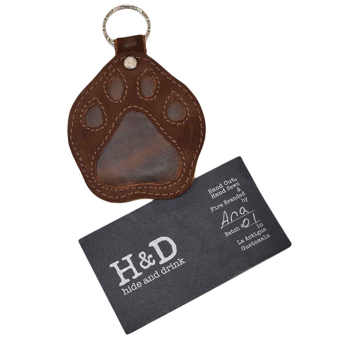 The Hide & Drink Doggy Paw Keychain displayed next to a business card, perfect for dog lovers. This artisan-made keychain showcases unique craftsmanship and full grain leather with tags like Hand Cut, Hand Sewn, and Handmade in La Antigua Guatemala & Oaxaca Mexico, making it a thoughtful gift option.
