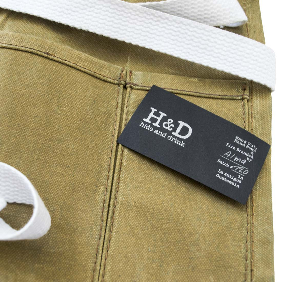 Close-up of the label on the Waxed Canvas Waist Apron by Hide & Drink, highlighting its commitment to small batch production and artisan craftsmanship. This apron combines water resistant waxed canvas with hand cut and hand sewn details, proudly made in La Antigua Guatemala & Oaxaca Mexico.
